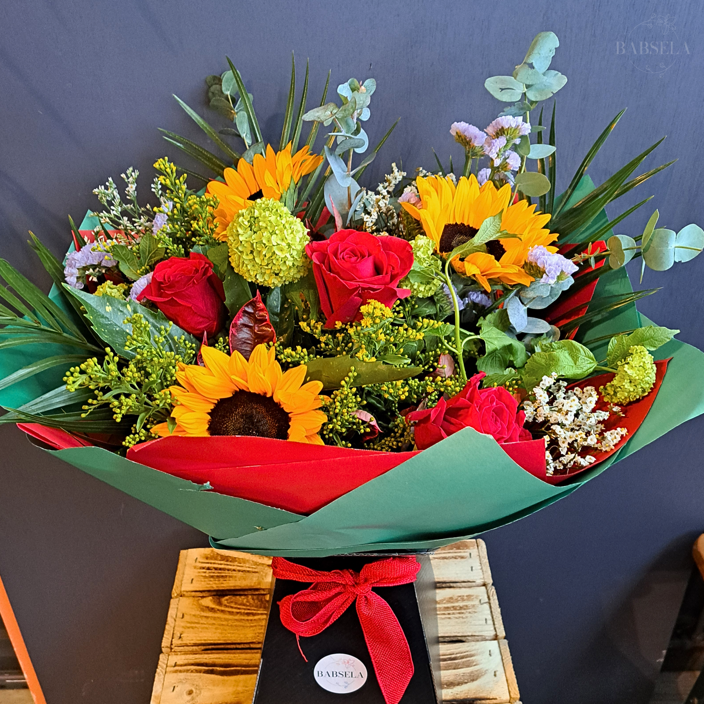 A vibrant bouquet featuring sunflowers, red roses, green hydrangeas, and assorted greenery, wrapped in red and green paper, displayed against a dark background