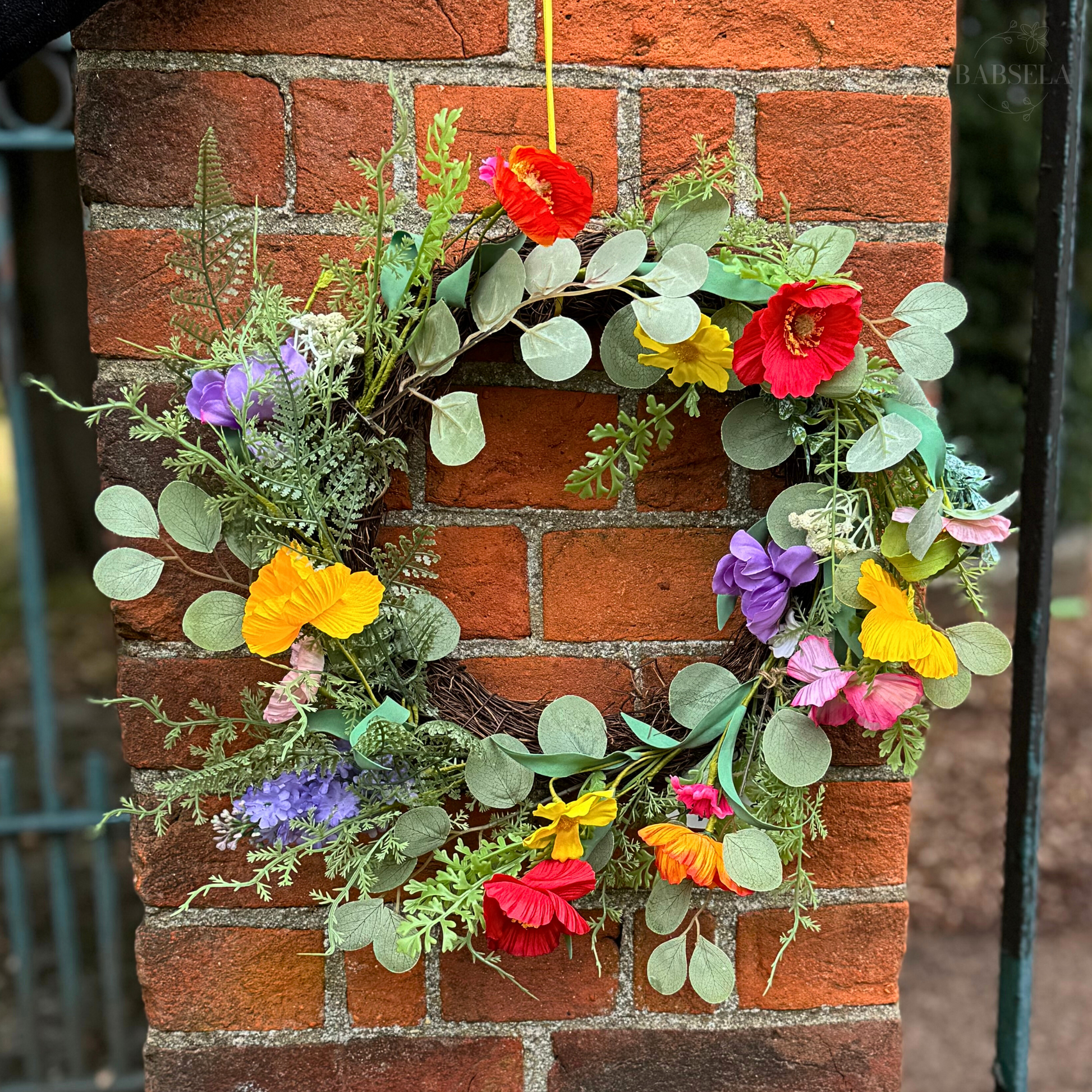 A vibrant floral wreath featuring colorful artificial flowers and green foliage, hanging on a red brick wall