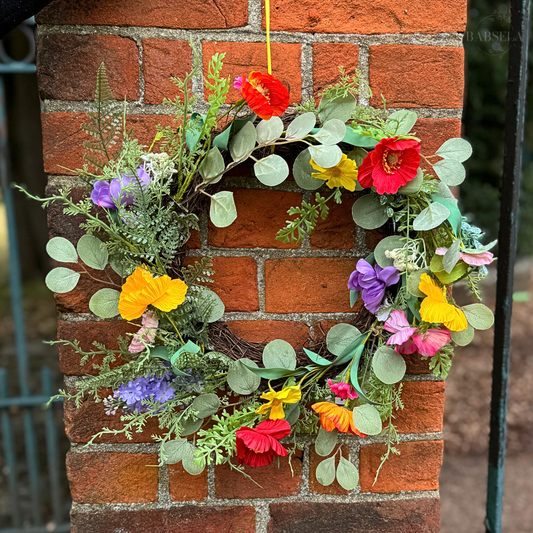 A vibrant floral wreath featuring colorful artificial flowers and green foliage, hanging on a red brick wall