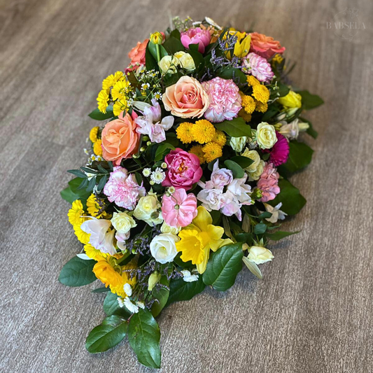 Elegant floral spray arrangement featuring a mix of roses, carnations, daisies, and yellow pom-poms in vibrant shades of pink, peach, yellow, and white, accented with lush green foliage